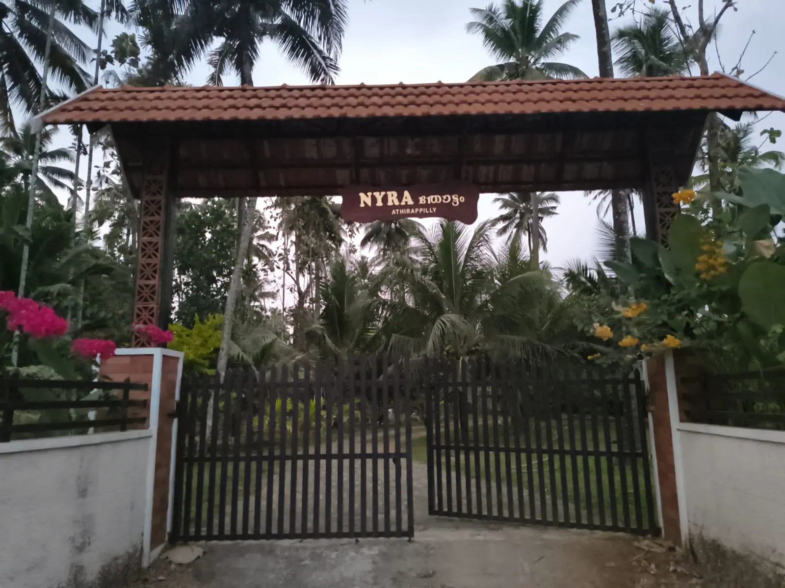 Tile-roofed entrance gateway of Nyra Thottam in Athirappilly with the property sign, flowering bougainvillea and coconut palms behind